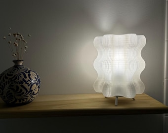 Op de afbeelding: Een witte tafellamp met een golvende, geplooide kap. De lamp staat op een houten plank met een blauw-witte gebloemde vaas met gedroogde bloemen.