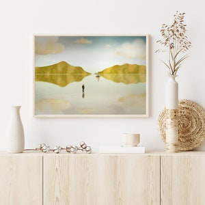 Puede incluir: Una impresi&oacute;n enmarcada de una pintura de paisaje minimalista con una sola figura de pie en el medio de una masa de agua reflectante. El agua refleja el cielo y dos colinas en la distancia. Un peque&ntilde;o velero se encuentra en la distancia.