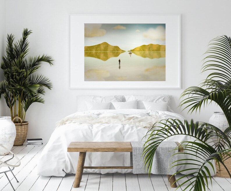 Puede incluir: Una cama blanca con ropa de cama blanca y un banco de madera delante. Una impresi&oacute;n enmarcada de un paisaje con un barco y una persona de pie en el agua cuelga de la pared detr&aacute;s de la cama. Una planta en maceta est&aacute; en el lado izquierdo de la imagen.