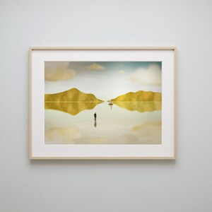 Puede incluir: Una impresi&oacute;n enmarcada de un paisaje surrealista con dos islas, un velero y una figura solitaria de pie en el agua. El cielo es de un azul p&aacute;lido con nubes blancas, y el agua es de un oro reflectante.