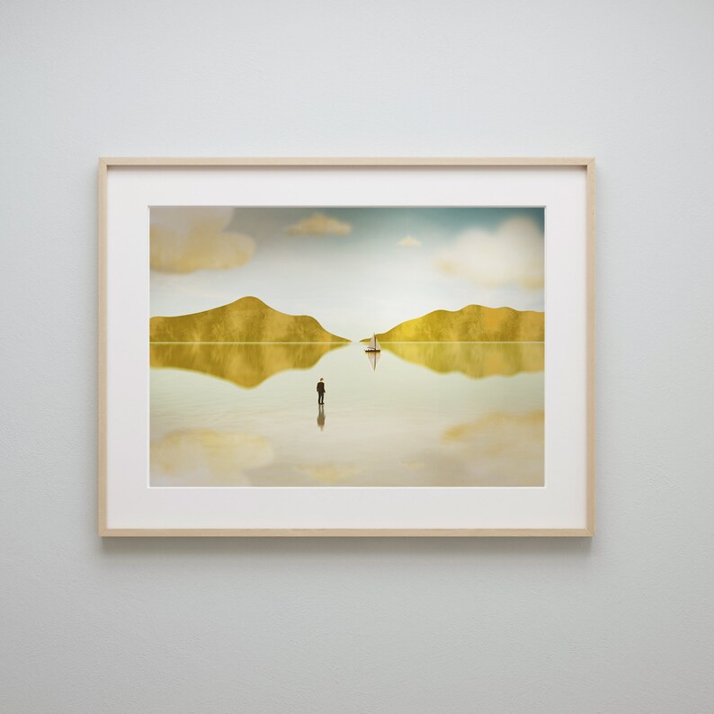 Puede incluir: Una impresi&oacute;n enmarcada de un paisaje surrealista con dos islas, un velero y una figura solitaria de pie en el agua. El cielo es de un azul p&aacute;lido con nubes blancas, y el agua es de un oro reflectante.