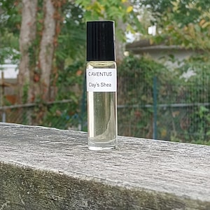 May include: A clear glass roll-on bottle with a black cap, filled with a light yellow liquid. The label reads "CAVENTUS Clay's Shea." The bottle is set against a weathered wooden surface with a blurred green background of trees and foliage.