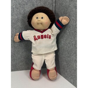 Può includere: Una bambola Cabbage Patch Kids con capelli in lana marrone scuro e viso rotondo. La bambola indossa una divisa da baseball color crema con la scritta rossa Angels sul petto e bordi a righe rosse e blu sulle maniche e sui pantaloni.