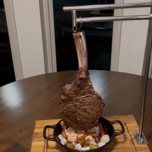 May include: A large, cooked tomahawk steak suspended above a cast iron dish filled with prawns, vegetables, and rice. The steak is attached to a metal stand. The dish sits on a wooden chopping board. The background is a dark, round table and a window.