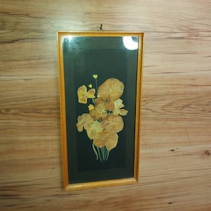 Könnte beinhalten: Ein gerahmtes Arrangement aus gepressten Blumen. Der rechteckige Rahmen hat einen hellbraunen Holzrahmen. Die Blumen sind hellbraun und vor einem schwarzen Hintergrund angeordnet. Der Rahmen hängt an einer Holzwand.