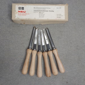 Op de afbeelding: Een set van zes houtdraaibeitels met houten handvatten, in een waaier gerangschikt. De beitels zijn zilverkleurig met houten handvatten. Een kartonnen doos met Duitse tekst staat op de achtergrond.