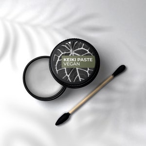 Puede incluir: Un tarro negro con un diseño de hoja blanca y el texto "KEIKI PASTE VEGAN" en la tapa. Un palo de madera con una punta negra está al lado del tarro.