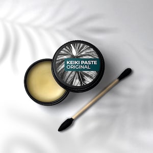 Könnte beinhalten: Ein schwarzes Glas mit einem weißen Etikett, auf dem "KEIKI PASTE ORIGINAL" und ein schwarz-weißes Blattdesign steht. Das Glas ist geöffnet und ein Holzstab mit schwarzer Spitze liegt daneben.
