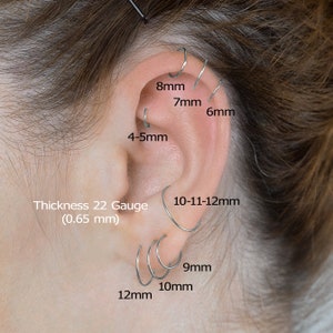May include: A close-up of a person's ear with multiple silver hoop earrings in various sizes. The earrings are labeled with their measurements in millimeters: 8mm, 7mm, 6mm, 4-5mm, 10-11-12mm, 9mm, 10mm, and 12mm. The text 'Thickness 22 Gauge (0.65 mm)' is also visible.