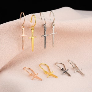 May include: Six pairs of sword-shaped dangle earrings in gold, silver, and rose gold. The earrings are on a pink fabric background.