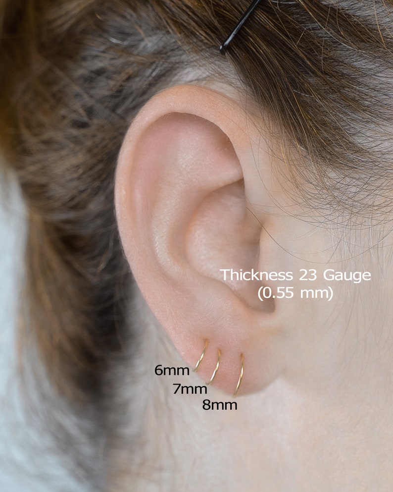 May include: Three gold hoop earrings in different sizes: 6mm, 7mm, and 8mm. The earrings are shown in a person's ear. The text 'Thickness 23 Gauge (0.55 mm)' is also visible.