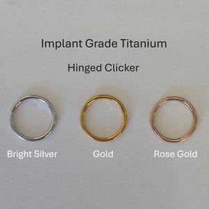 May include: Three hinged clicker rings in bright silver, gold, and rose gold. The text reads "Implant Grade Titanium" and "Hinged Clicker". These rings are designed for body piercings.