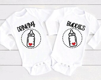 May include: Two white long-sleeve baby bodysuits featuring black text and graphics. The left bodysuit displays the word DRINKING above a baby bottle icon with a red heart. The right bodysuit displays the word BUDDIES above a matching bottle icon.