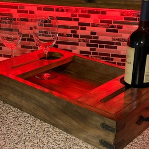 May include: A wooden wine serving tray with two wine glasses and a wine bottle. The tray has cutouts for the glasses and bottle, and a handle. The tray is stained a dark brown colour and has metal accents.
