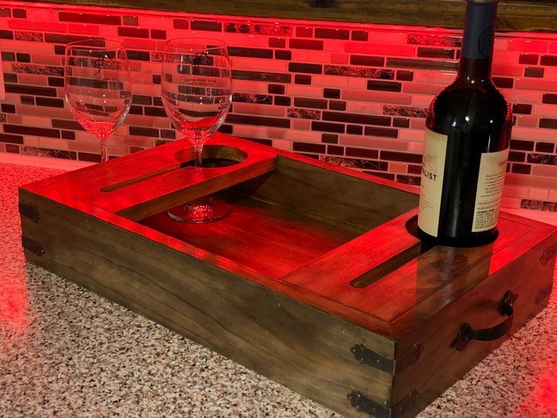 May include: A wooden wine serving tray with two wine glasses and a wine bottle. The tray has cutouts for the glasses and bottle, and a handle. The tray is stained a dark brown colour and has metal accents.