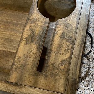 May include: Wooden wine bottle holder with a distressed finish. The holder has a circular opening for the bottle and a slot for the neck. The surface is decorated with vintage-style text and graphics. A black handle is attached to the side.