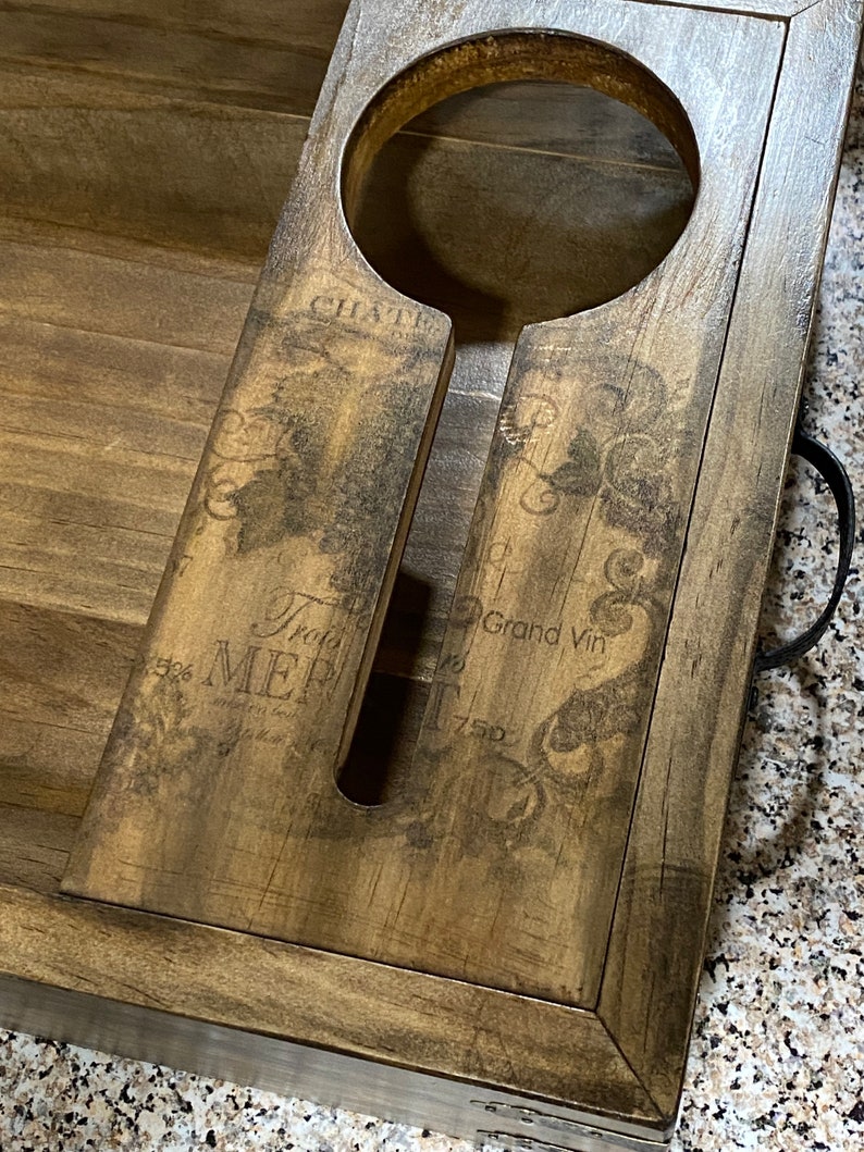 May include: Wooden wine bottle holder with a distressed finish. The holder has a circular opening for the bottle and a slot for the neck. The surface is decorated with vintage-style text and graphics. A black handle is attached to the side.