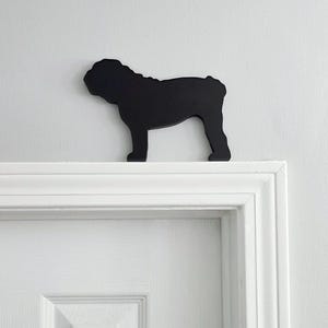 May include: A solid black silhouette of a bulldog standing in profile. The cutout figure features the distinct stocky build, wrinkled head, and short tail characteristic of the breed, presented as a flat, matte decorative object.
