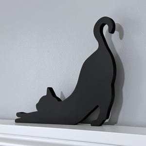 A black wooden cat silhouette wall decor. The cat is in a stretched out pose with its tail curled up. The cat is facing to the left.