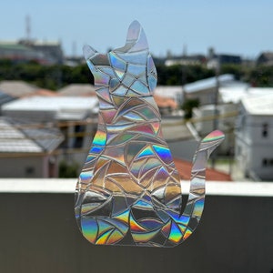 May include: A cat-shaped window cling with a mosaic-like design. The cling is iridescent, reflecting a spectrum of colors including blues, greens, and yellows. The cat is in a sitting position, with its tail curled.