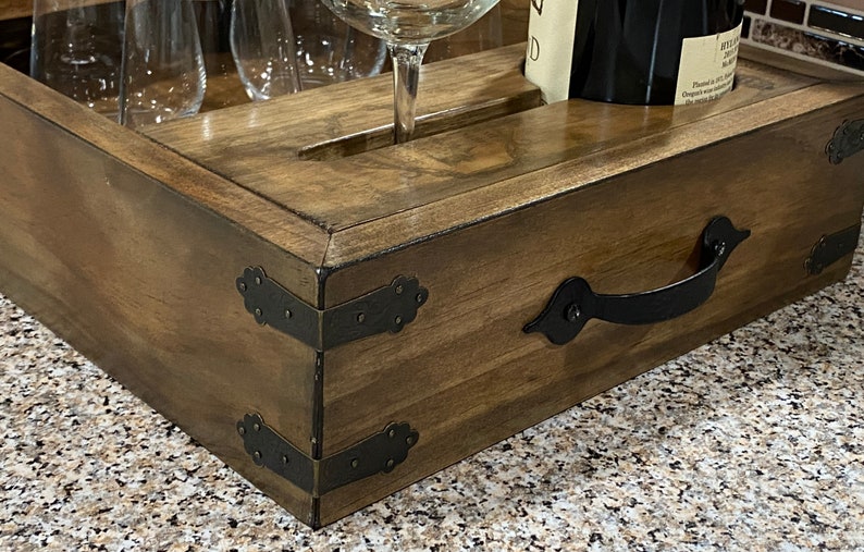 May include: Wooden wine serving tray with a dark stain, featuring a handle and decorative metal corner accents. The tray has slots for wine glasses and a bottle, ideal for serving.