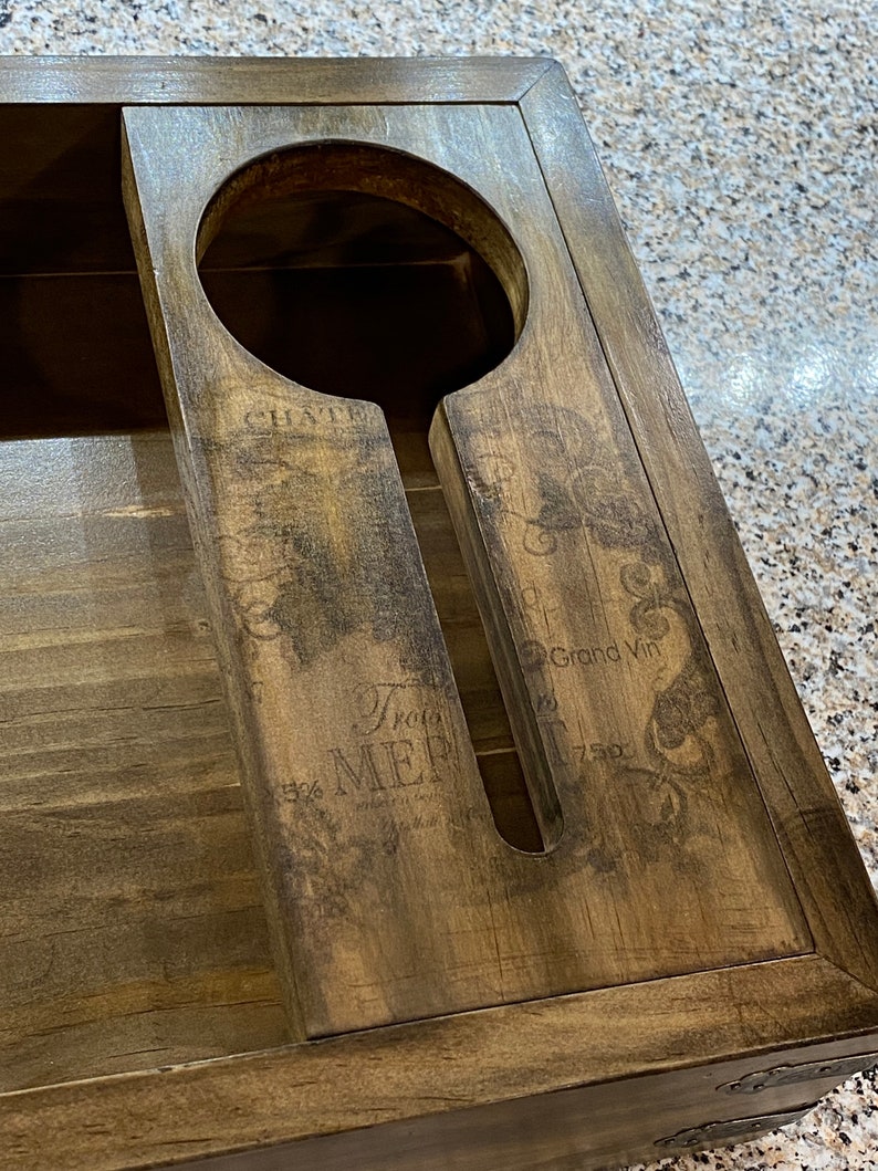 May include: A dark brown wooden wine bottle and glass holder. It has a circular space for a bottle and a slot for a glass. The surface is decorated with printed text and ornamental designs.