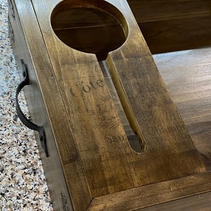 May include: A dark-stained wooden wine serving tray. It features a circular bottle holder and a slot for a glass. Metal corner brackets and a handle are attached. The word "Cote" is subtly visible on the wood's surface.