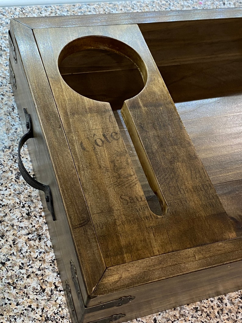 May include: A dark-stained wooden wine serving tray. It features a circular bottle holder and a slot for a glass. Metal corner brackets and a handle are attached. The word "Cote" is subtly visible on the wood's surface.