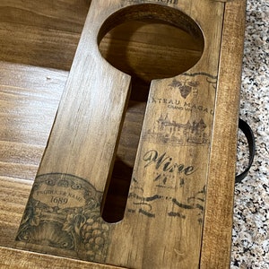May include: Wooden wine serving tray with a handle. The tray has a cutout for a wine glass and is decorated with wine-themed graphics and text, including "MAISON BLANCHE" and "Wine". The tray is made of light brown wood.