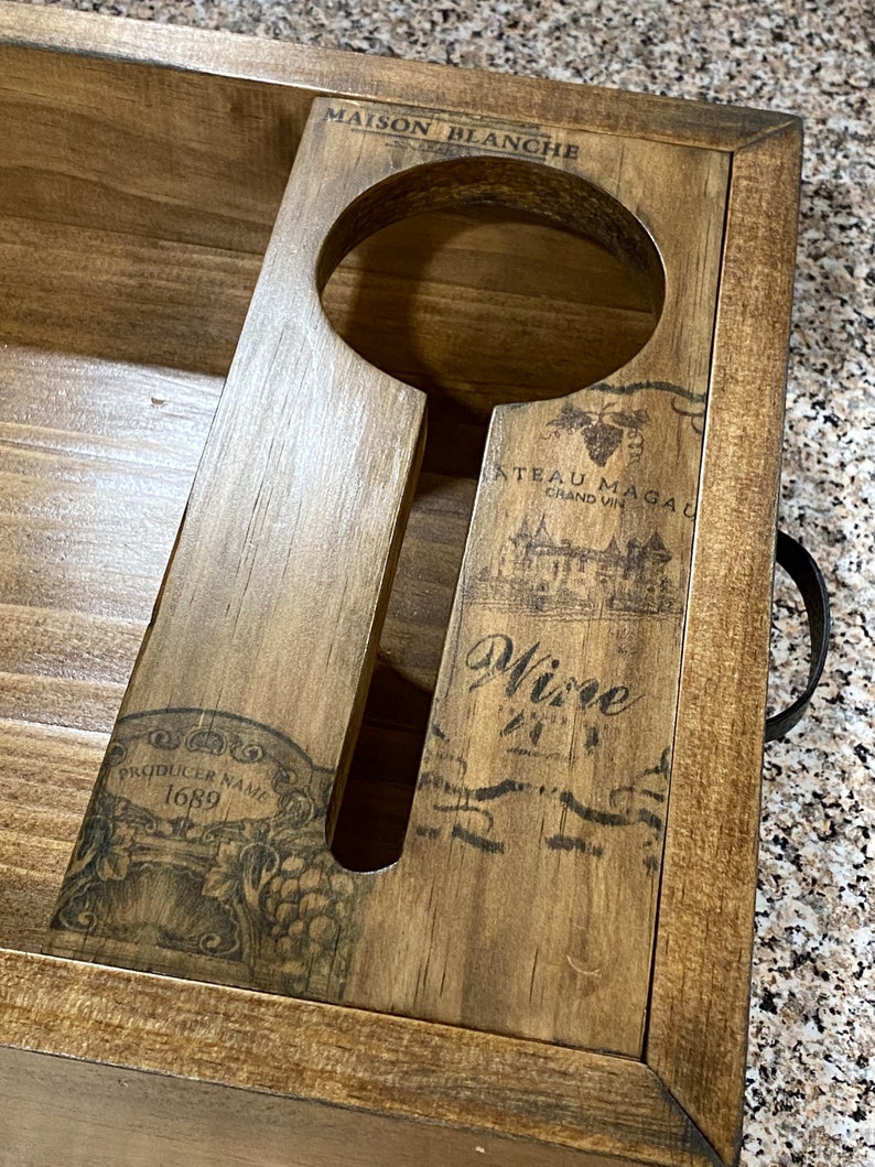 May include: Wooden wine serving tray with a handle. The tray has a cutout for a wine glass and is decorated with wine-themed graphics and text, including "MAISON BLANCHE" and "Wine". The tray is made of light brown wood.