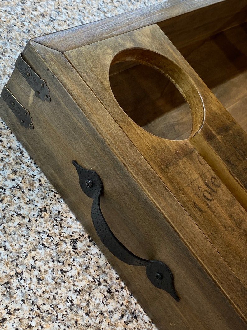 May include: A wooden wine bottle holder with a dark stain. It features a circular cutout for the bottle and a handle. The holder has decorative metal corner brackets and a handle. The word "Cote" is faintly visible.