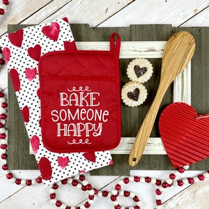May include: A red quilted pot holder with the white text BAKE SOMEONE HAPPY, a white kitchen towel with red hearts, a wooden spoon, two heart-shaped cookies, a red corrugated heart ornament, and a string of red and white wooden beads.