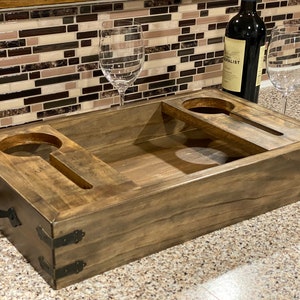 May include: A wooden wine serving tray with two circular cutouts for wine glasses and two slots for holding wine glasses. The tray is stained a dark brown and has metal corner accents. A bottle of wine and two wine glasses are on the tray.