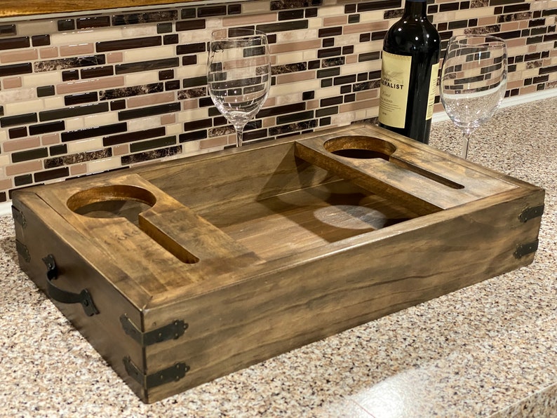 May include: A wooden wine serving tray with two circular cutouts for wine glasses and two slots for holding wine glasses. The tray is stained a dark brown and has metal corner accents. A bottle of wine and two wine glasses are on the tray.