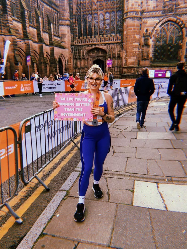 May include: A woman in blue athletic wear holds a pink sign with the text "GO LIV! YOU'RE RUNNING BETTER THAN THE GOVERNMENT." She wears glasses and a medal. The background features a historic building and a running event.
