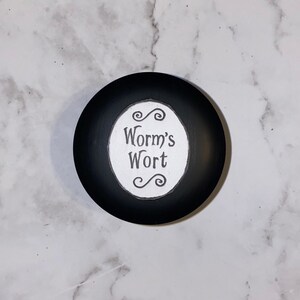 Può includere: Un contenitore nero e rotondo con la scritta "Worm's Wort" in un ovale bianco. Il testo è in un carattere stravagante, con riccioli decorativi sopra e sotto. Probabilmente un coperchio o un piccolo barattolo.