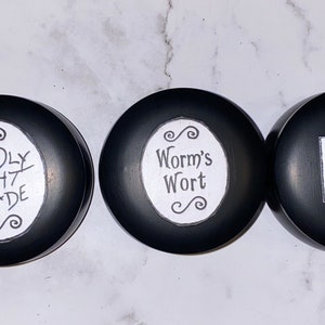 Può includere: Tre contenitori rotondi neri con etichette bianche. Le etichette riportano il seguente testo: "Deadly Nightshade", "Worm's Wort" e "Frog's Breath".