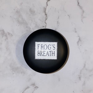 May include: A black round candle jar with a white label that reads "FROG'S BREATH".