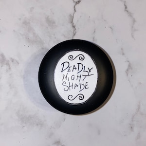 Può includere: Un contenitore rotondo nero con un'etichetta bianca su cui è scritto "DEADLY NIGHT SHADE" in corsivo nero.