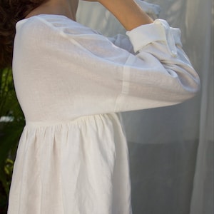 Puede incluir: Vestido de lino blanco con mangas largas y cintura fruncida. El vestido presenta un cuello redondo y pu&ntilde;os abotonados. La tela parece ligera y transpirable, adecuada para climas c&aacute;lidos.