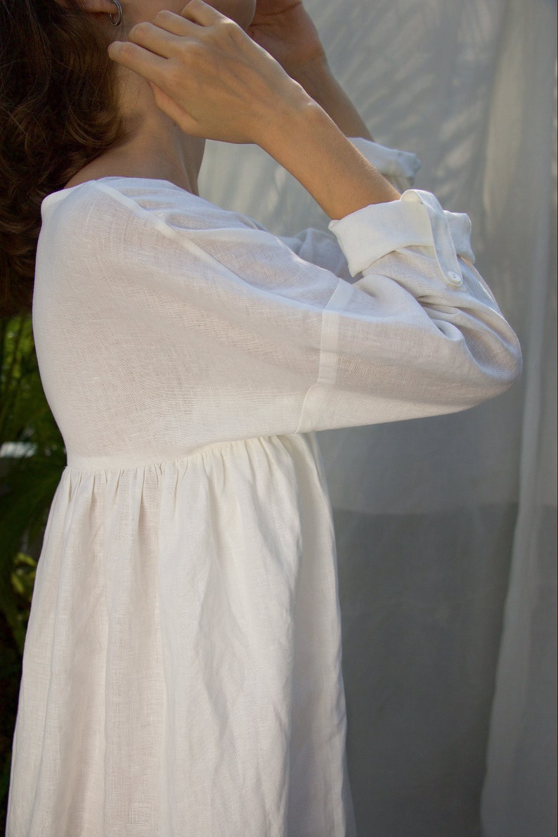 Puede incluir: Vestido de lino blanco con mangas largas y cintura fruncida. El vestido presenta un cuello redondo y pu&ntilde;os abotonados. La tela parece ligera y transpirable, adecuada para climas c&aacute;lidos.