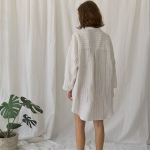 K&ouml;nnte beinhalten: Ein wei&szlig;es Leinenhemdkleid mit d&uuml;nnen schwarzen Streifen. Das Kleid hat einen Kragen und lange &Auml;rmel. Das Model tr&auml;gt das Kleid und steht auf einer wei&szlig;en Oberfl&auml;che.