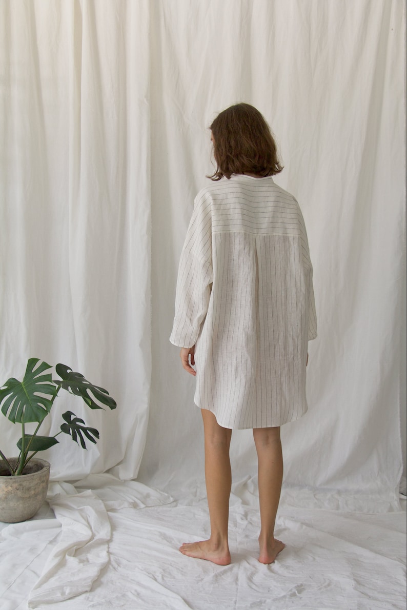 K&ouml;nnte beinhalten: Ein wei&szlig;es Leinenhemdkleid mit d&uuml;nnen schwarzen Streifen. Das Kleid hat einen Kragen und lange &Auml;rmel. Das Model tr&auml;gt das Kleid und steht auf einer wei&szlig;en Oberfl&auml;che.
