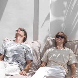 May include: Two people wearing sunglasses are relaxing on beige bean bag chairs. The person on the left is wearing a light gray t-shirt and white pants. The person on the right is wearing a light beige t-shirt and white pants.