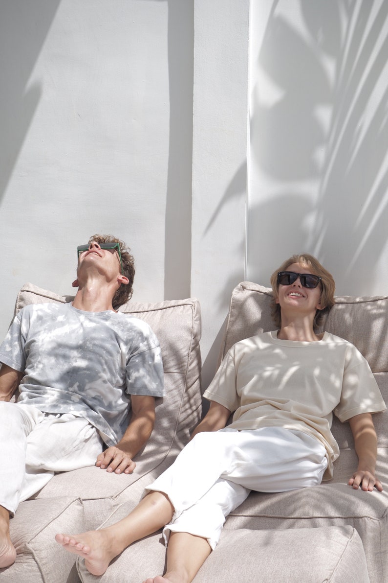 May include: Two people wearing sunglasses are relaxing on beige bean bag chairs. The person on the left is wearing a light gray t-shirt and white pants. The person on the right is wearing a light beige t-shirt and white pants.