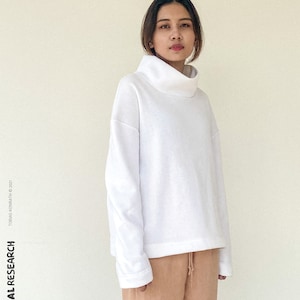 May include: A person wearing a white turtleneck sweatshirt and tan drawstring trousers. The sweatshirt has a wide, folded collar and long sleeves. The text "the SWEAT-SHIRT" is in the upper right corner. The text "TROPICAL RESEARCH" is on the left side.