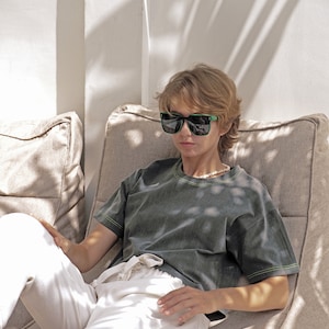 May include: A person wearing black sunglasses and a dark green t-shirt with a light green trim sits on a beige couch. The person is wearing white pants and has short blonde hair.