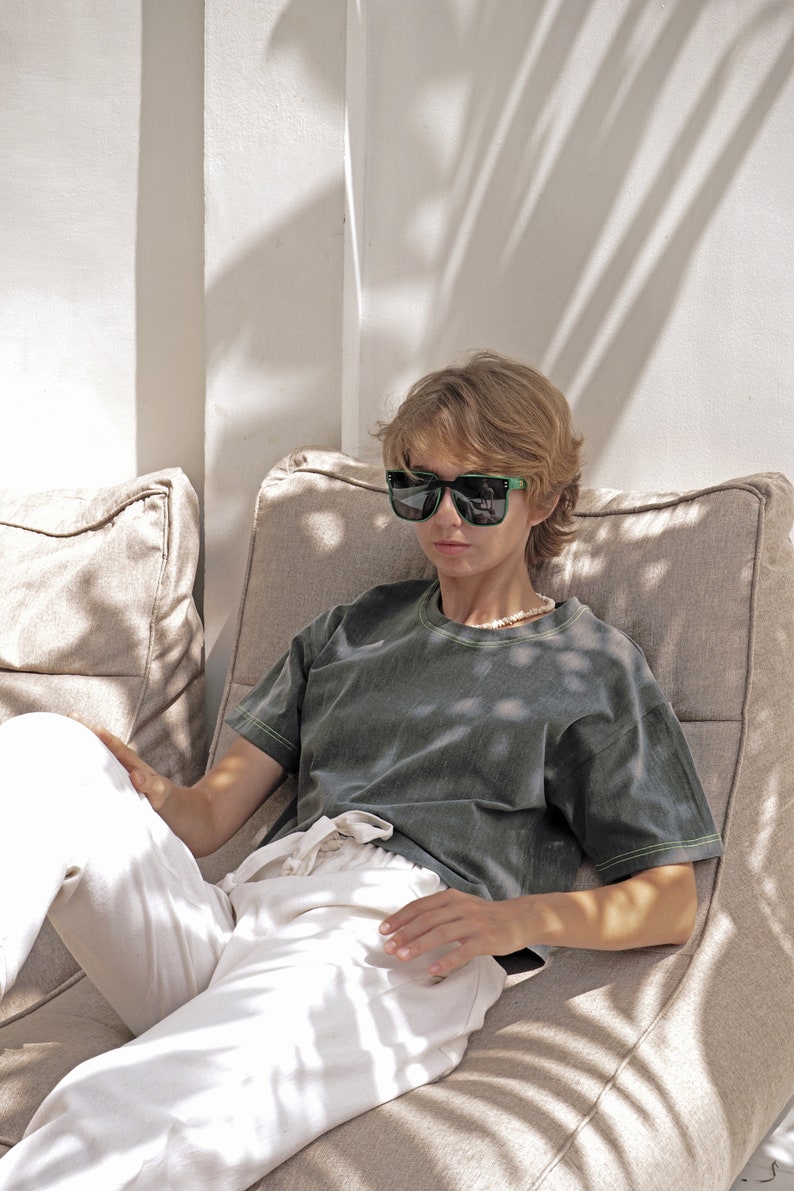 May include: A person wearing black sunglasses and a dark green t-shirt with a light green trim sits on a beige couch. The person is wearing white pants and has short blonde hair.
