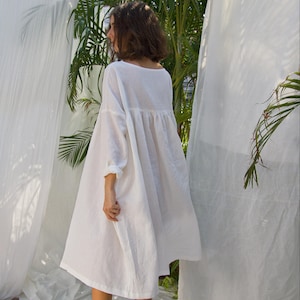 Puede incluir: Vestido de lino blanco con una silueta suelta y fluida. El vestido presenta un escote redondo, mangas largas y cintura fruncida. La modelo est&aacute; de pie descalza sobre una s&aacute;bana blanca, con cortinas blancas y follaje verde al fondo.