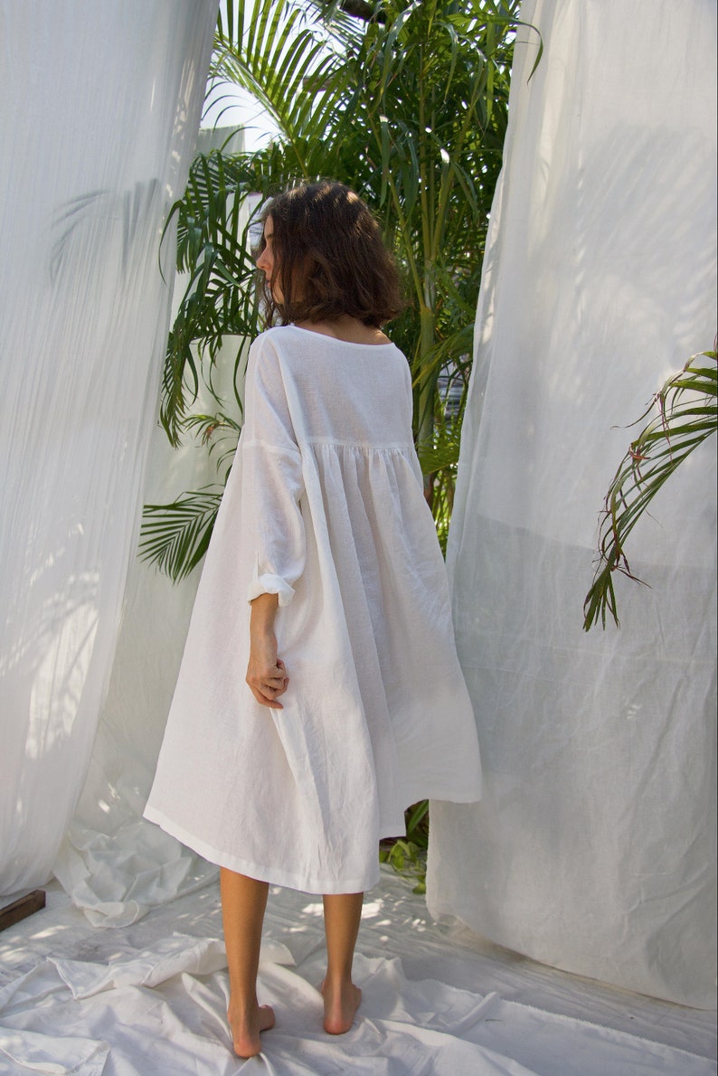 Puede incluir: Vestido de lino blanco con una silueta suelta y fluida. El vestido presenta un escote redondo, mangas largas y cintura fruncida. La modelo est&aacute; de pie descalza sobre una s&aacute;bana blanca, con cortinas blancas y follaje verde al fondo.